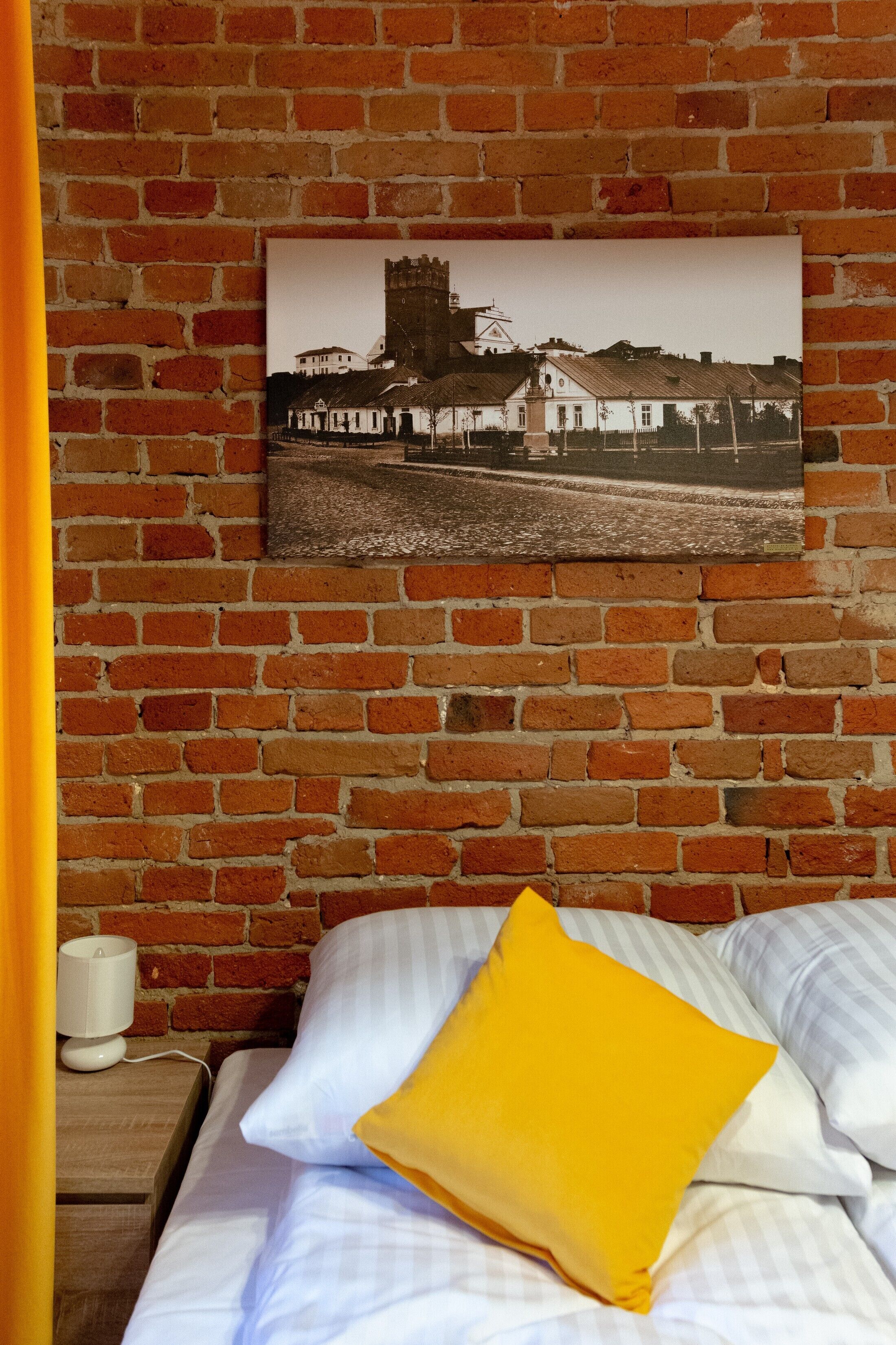 Un lit avec une literie blanche et à rayures, un oreiller jaune et une photographie en noir et blanc encadrée représentant un bâtiment sur un mur de briques.