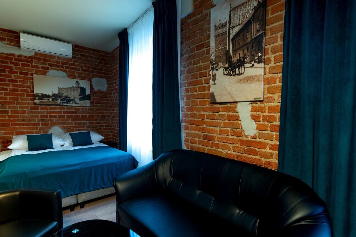 A hotel room with a brick wall, a bed with green bedding, a black leather sofa, and a large window with curtains.