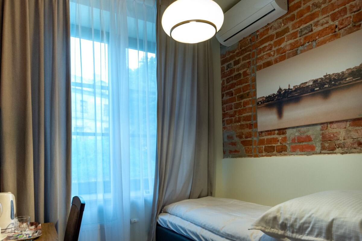 A compact room with a bed, a wooden table, a chair, a hanging light fixture, and a brick wall with an air conditioning unit.
