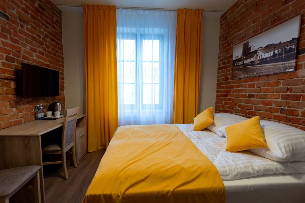 A hotel room with a brick wall, a bed with yellow bedding, a desk with a chair, a television, and a window with curtains.