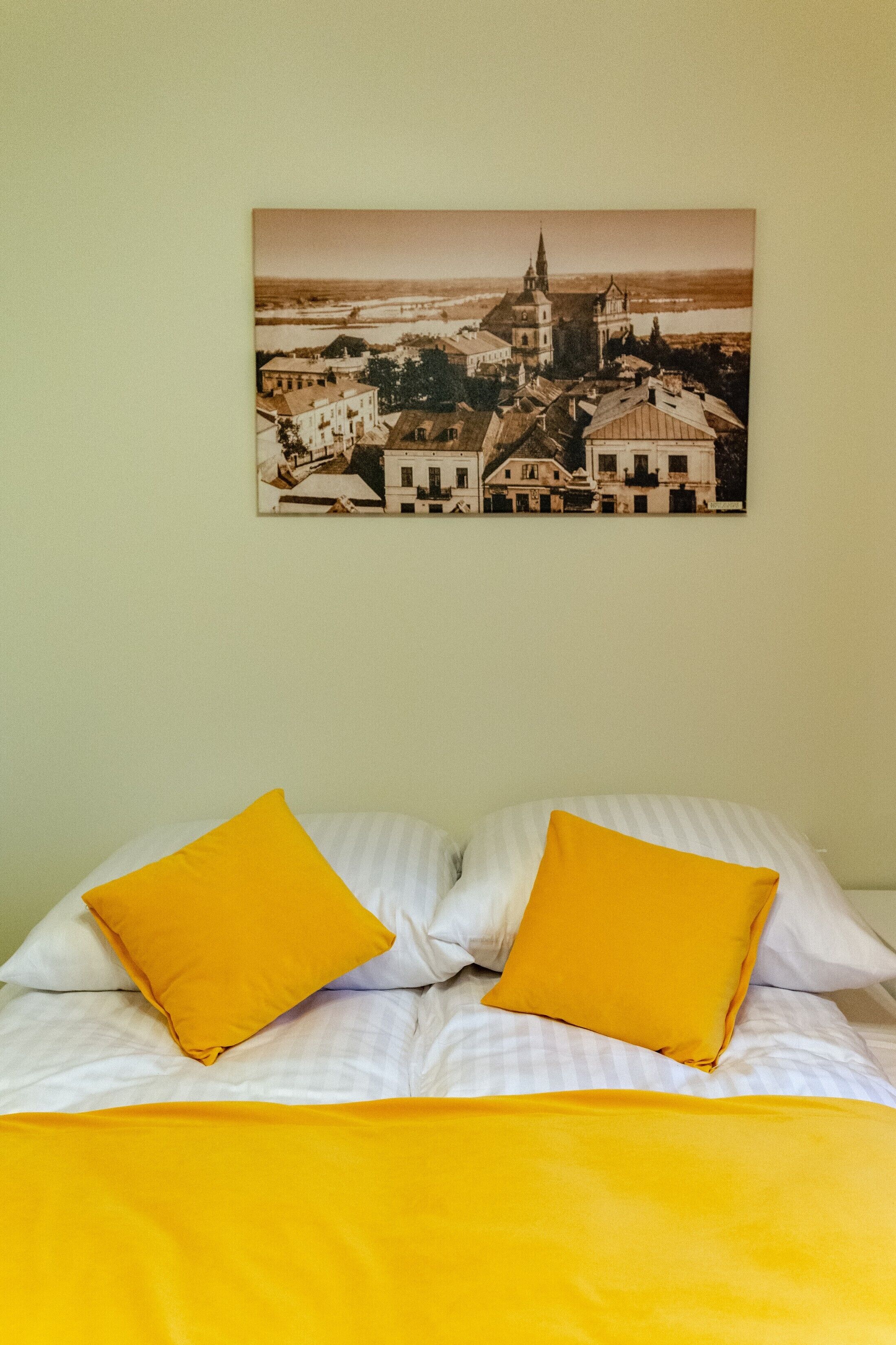 Un lit avec une literie blanche et deux coussins jaune vif, et au-dessus, un tableau encadré représentant une ville.
