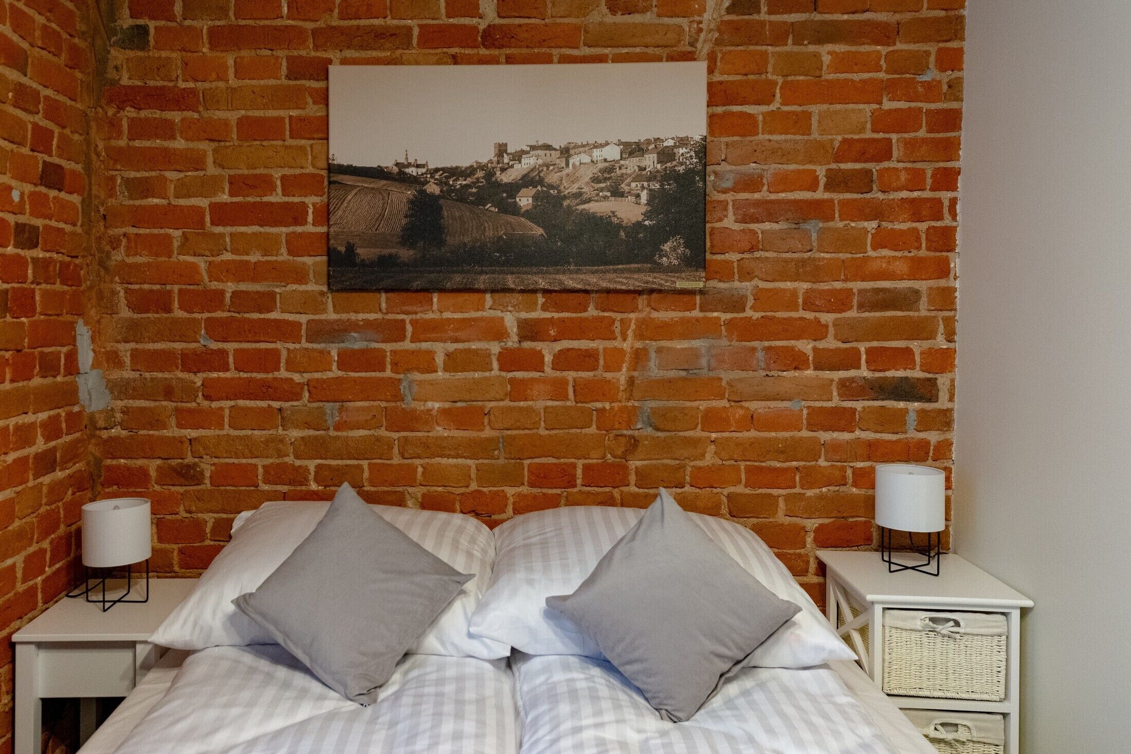 Una habitación con pared de ladrillo, cama con ropa de cama blanca y almohadas grises, mesita de noche con una lámpara y un marco de foto.