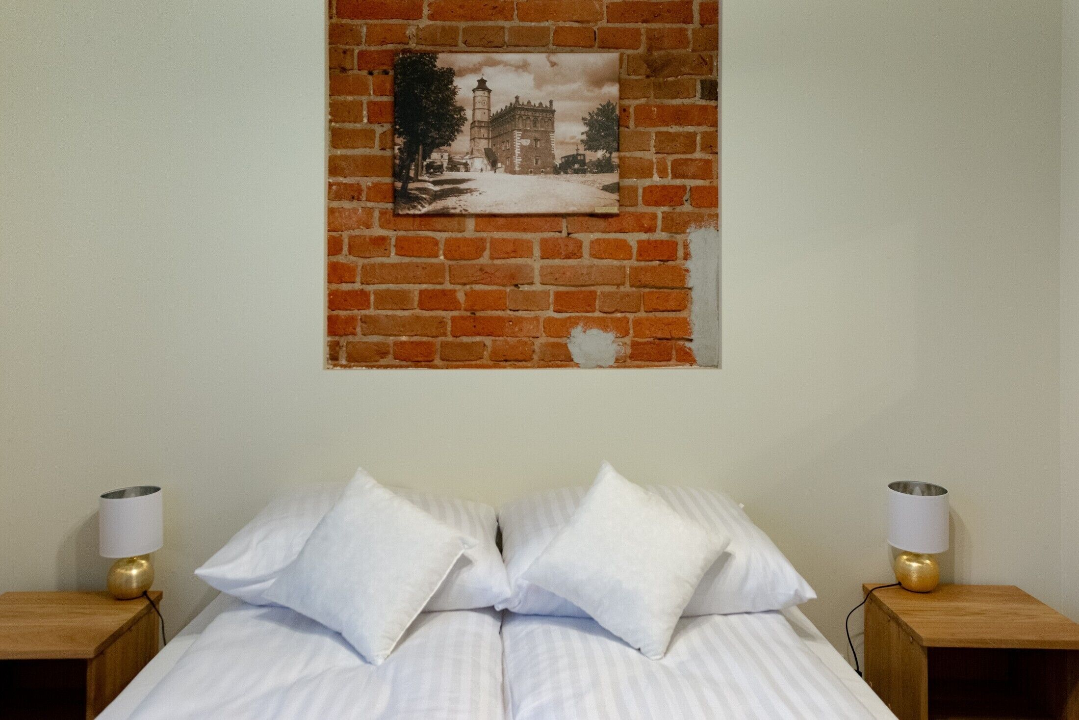 Una cama con ropa de cama blanca y dos almohadas, flanqueada por mesitas de noche de madera con lámparas, frente a una pared de ladrillo con un cuadro enmarcado en la parte superior.