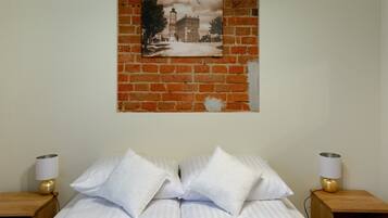 Una cama con ropa de cama blanca y dos almohadas, flanqueada por mesitas de noche de madera con lámparas, frente a una pared de ladrillo con un cuadro enmarcado en la parte superior.