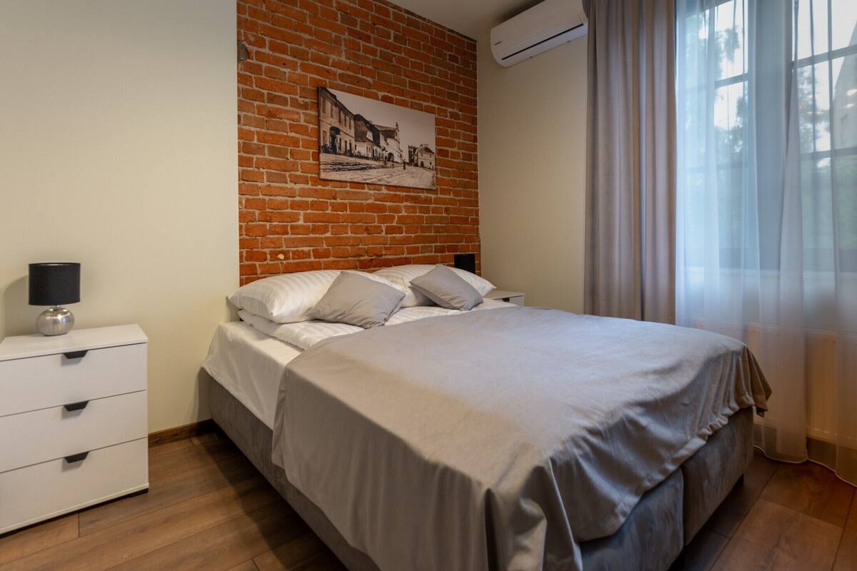 A bedroom with a brick wall, a bed with white bedding, a nightstand with a lamp, and a window with sheer curtains.