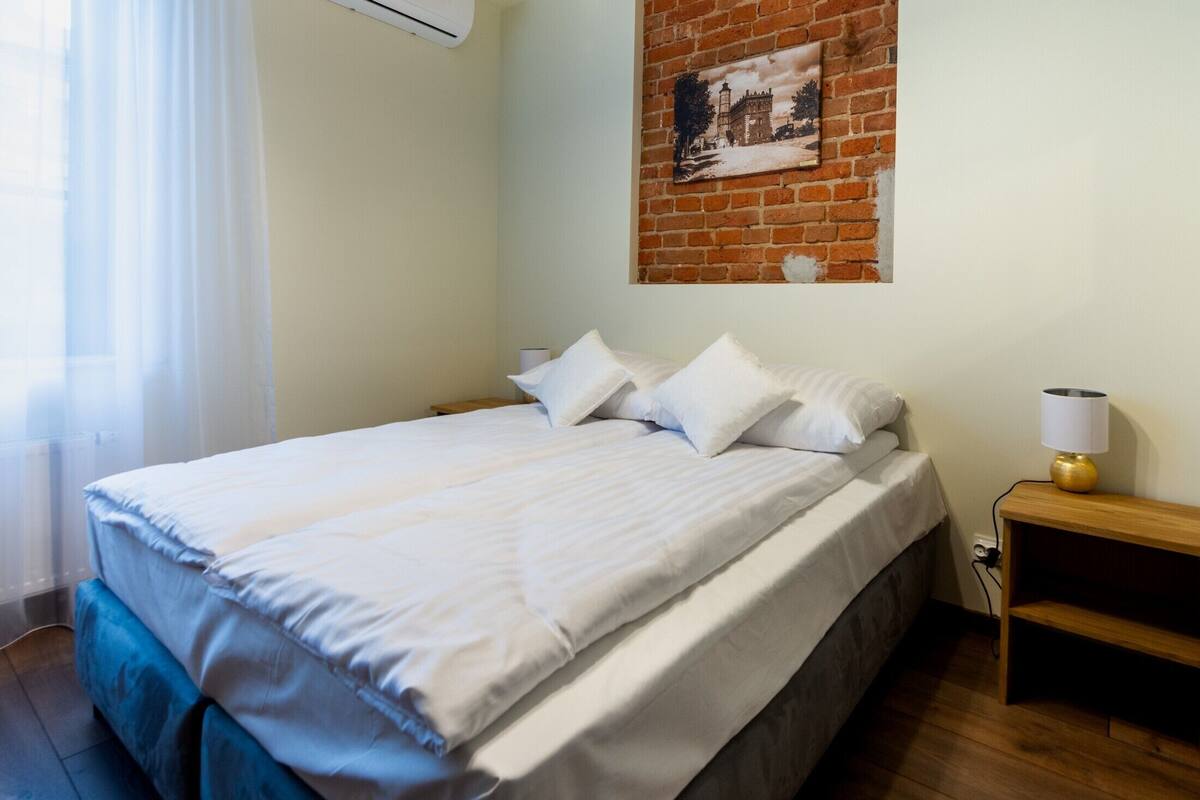 A neatly made bed with white linens, a wooden nightstand with a lamp, and a brick wall with a framed picture.