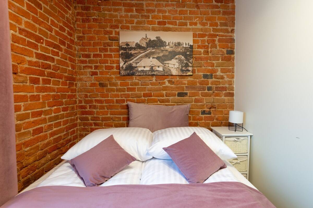 A bedroom with a brick wall, a bed with white and purple bedding, a nightstand with a lamp, and a framed picture on the wall.