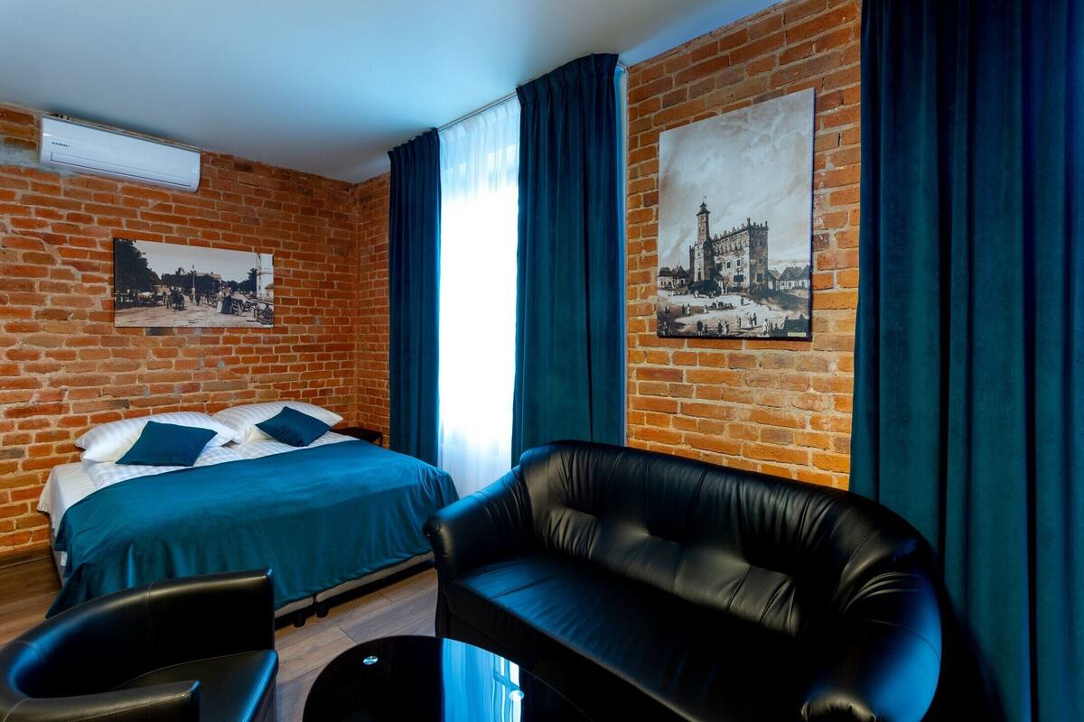 A hotel room with a brick wall, a bed with blue bedding, a black leather sofa, and two chairs.