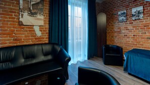 Une pièce avec un mur en briques, une grande fenêtre ornée de rideaux légers, un canapé en cuir noir, une petite chaise noire et un lit recouvert d’une couverture bleue.