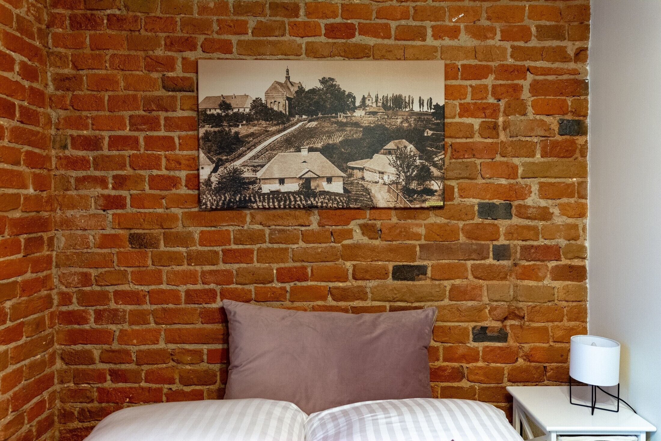 A room with a brick wall, a bed with a pillow, a small table with a lamp, and a framed picture of a landscape.