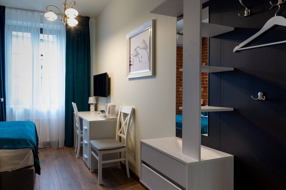 A compact bedroom with a bed, a desk, and a chair. There is a window with curtains, a wall-mounted TV, and a framed picture.