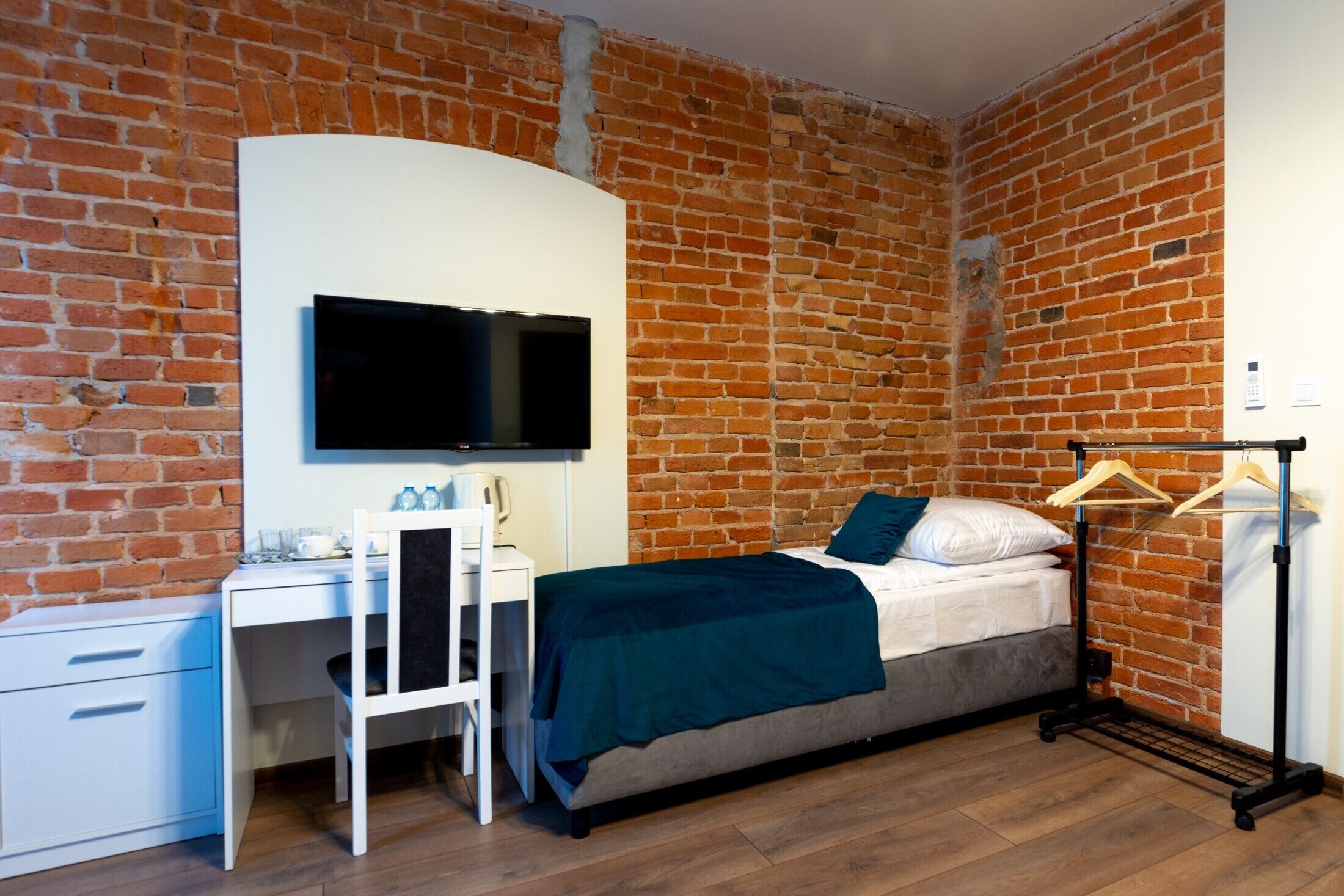 A bedroom with a brick wall, a bed, a desk with a chair, a TV, and a coat rack.