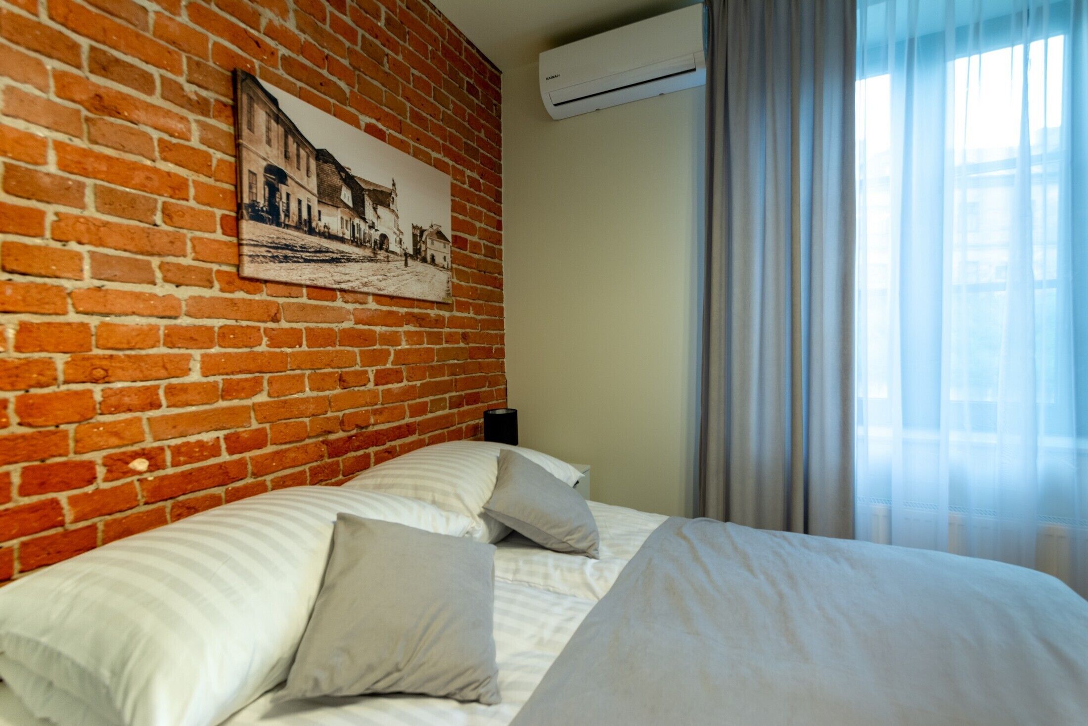 Una habitación de hotel con pared de ladrillo, cama con ropa blanca, un cuadro enmarcado y un equipo de aire acondicionado.
