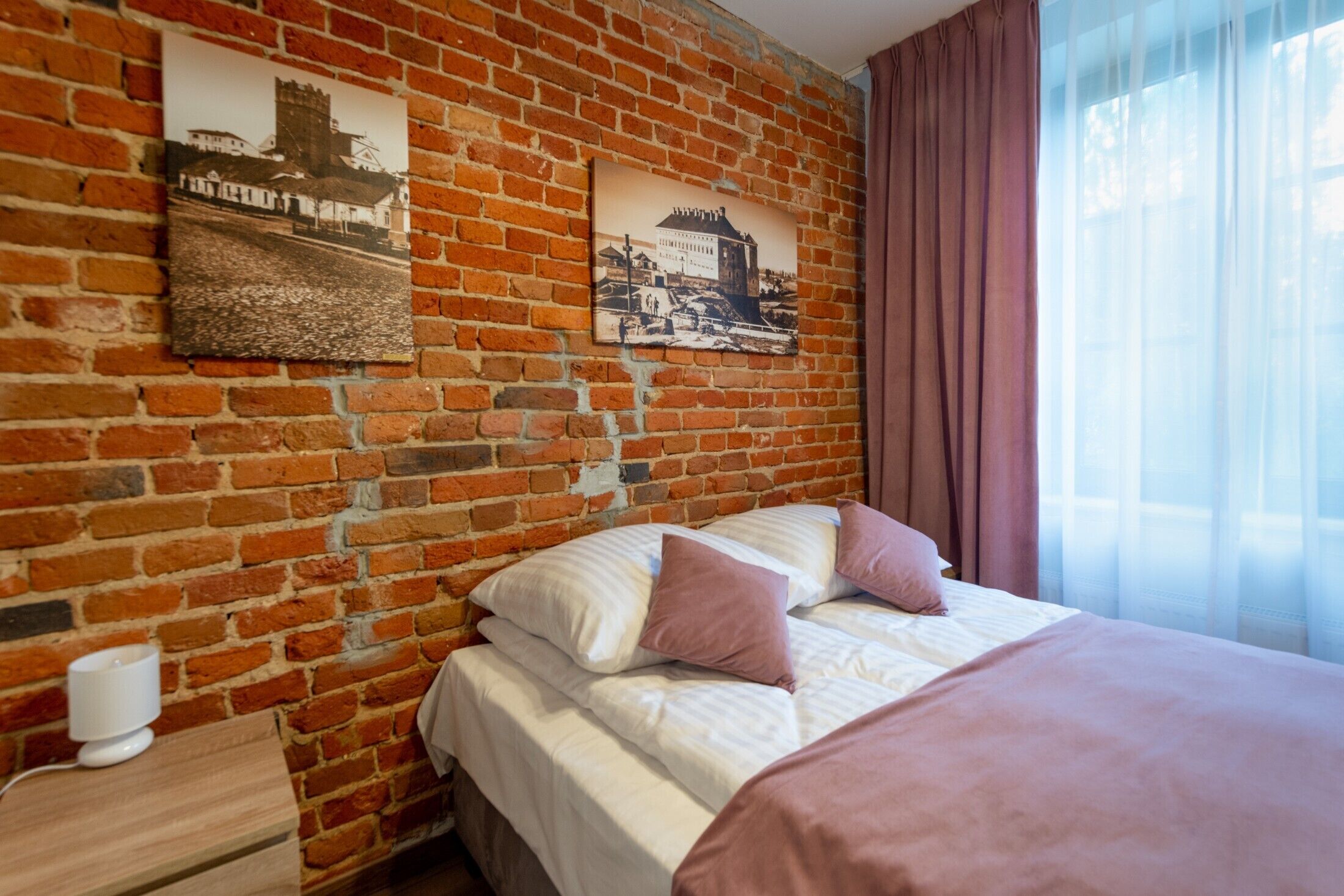 A hotel room with a brick wall, a bed with white and pink bedding, a nightstand with a lamp, and two framed pictures.