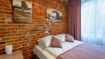 A hotel room with a brick wall, a bed with white and pink bedding, a nightstand with a lamp, and two framed pictures.