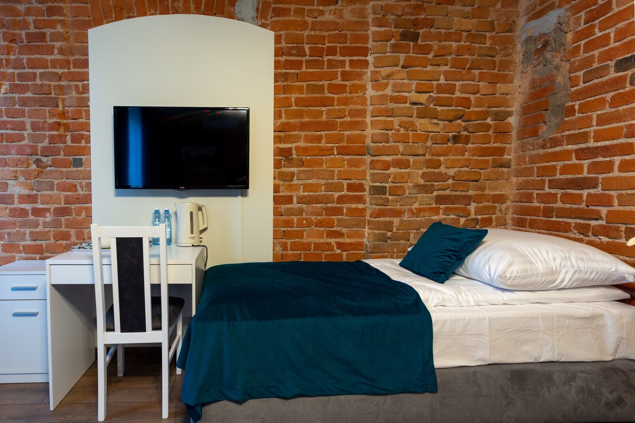 Quarto com parede de tijolos, cama com roupa de cama branca e manta verde-água, escrivaninha com cadeira e televisão de tela plana fixada acima da escrivaninha.