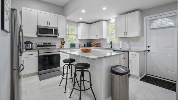 A modern kitchen with white cabinets, a central island with stools, stainless steel appliances, and a large window providing natural light. - Flexible Relocation Stay: Spacious, Pet-Friendly, w/ Utilities near Mid/Downtown (Atlanta)