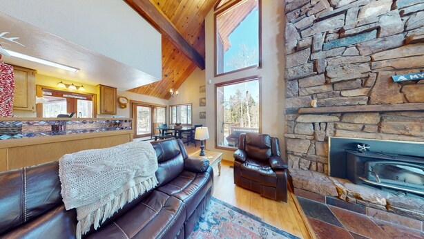 A cozy living room with a stone fireplace, leather sofa, and a large window overlooking a deck. - Large Deck | Fireplace | Forest Views | Kings Landing (Angel Fire)