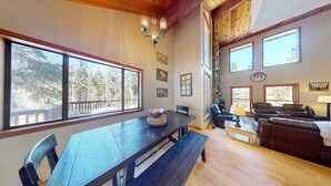 A spacious living room with a large wooden dining table, leather sofas, and multiple windows offering views of a wooded area. - Large Deck | Fireplace | Forest Views | Kings Landing (Angel Fire)