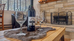Une bouteille de vin, deux verres et un tapis en fourrure sur une table en bois, dans une pièce dotée d’une cheminée en pierre et d’une fenêtre avec des stores.