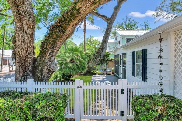 A white house with a white picket fence, a large tree, and a small garden with a pathway.