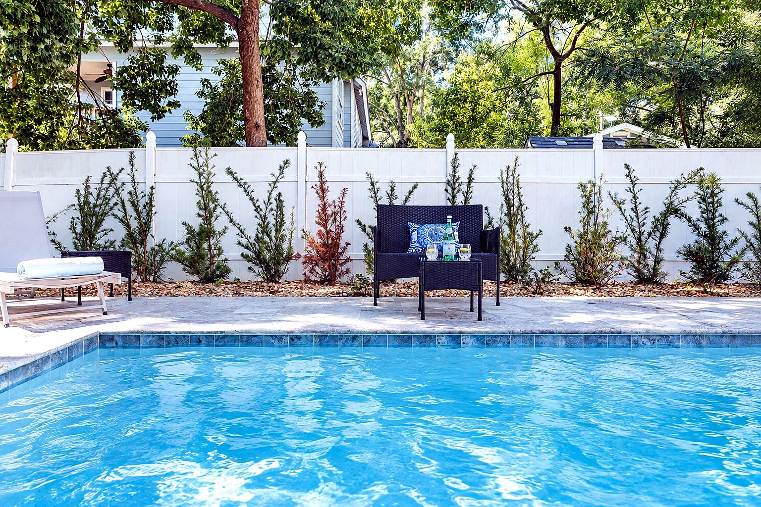 A swimming pool with a white fence, a lounge chair, and a table with a bottle.