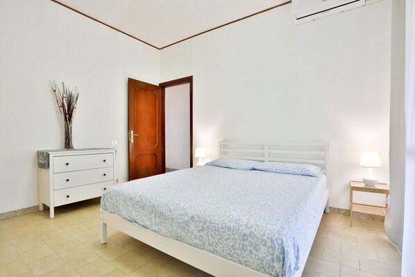 A bedroom with a bed, a dresser, a door, a bedside table with a lamp, and a wall-mounted air conditioner.