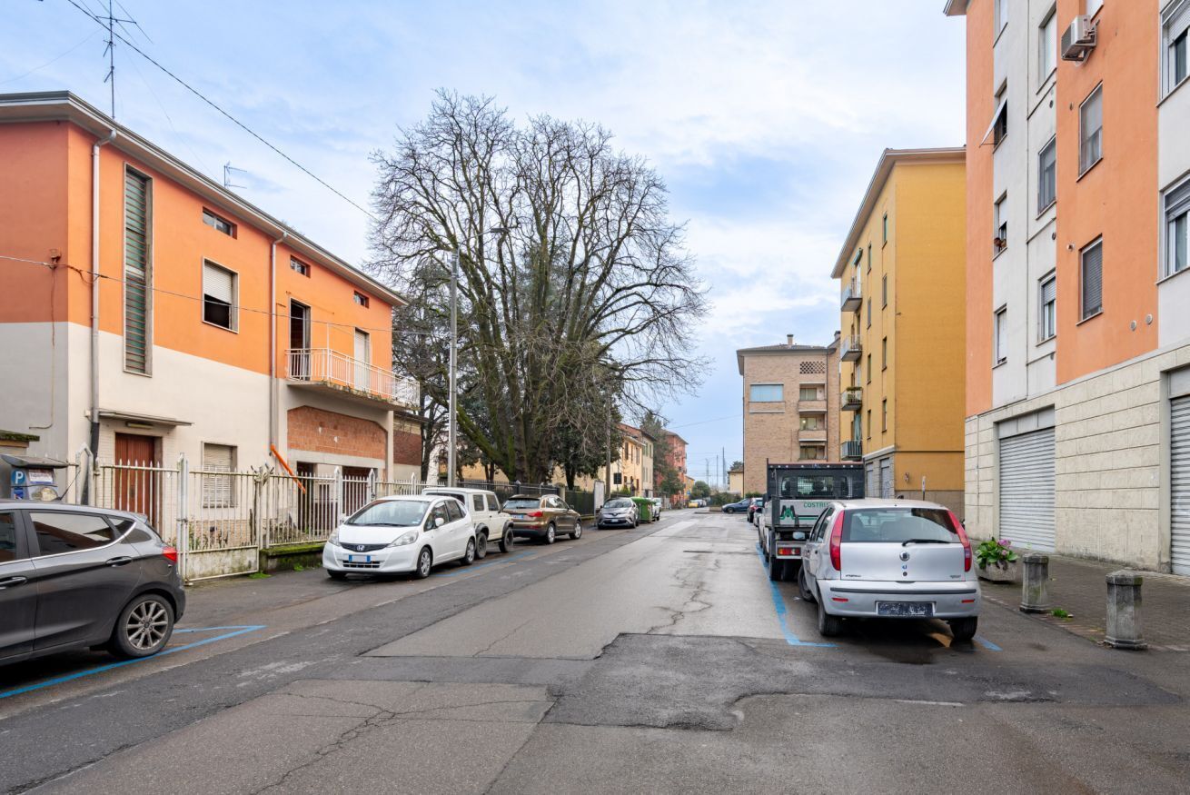A residential street with parked cars, apartment buildings, and a tree.