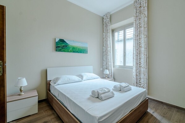 Un dormitorio con cama, mesita de noche, lámpara, cuadro y ventana con cortinas.
