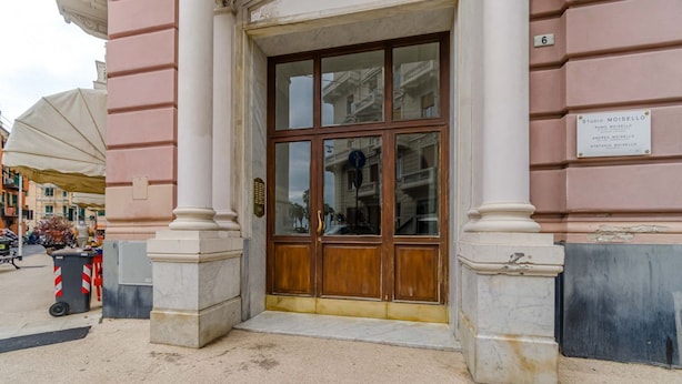 L'ingresso di un edificio con una grande porta in legno, una targa sopra e una vista della strada con auto parcheggiate e un bidone dell'immondizia.