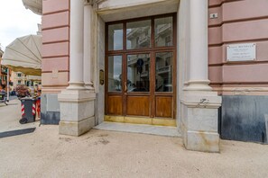 A building entrance with a large wooden door, a plaque above it, and a street view with parked cars and a trash bin.