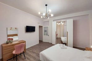 A modern hotel room with a wooden desk, a pink chair, a bed, a TV, and a mirror.