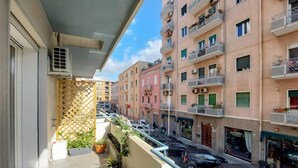 Un balcón con macetas, un cerramiento de celosía y vistas a una calle con coches aparcados y edificios de varios pisos.
