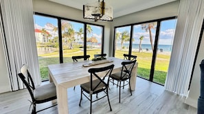 A dining area with a table and chairs, large windows with a view of a beach and palm trees.