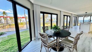 A spacious dining area with a round table and wicker chairs, large windows offering a view of a pool and outdoor area, and a kitchen area with a sink and countertop.