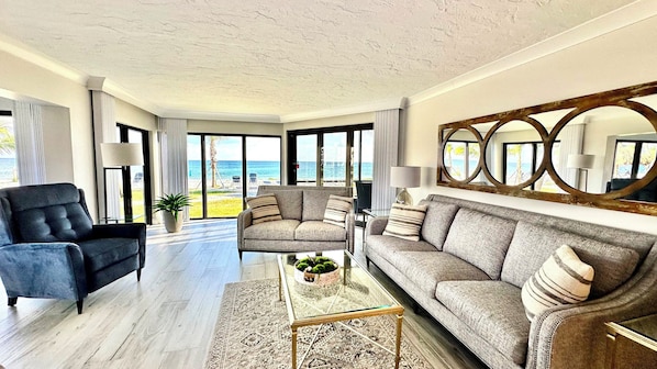 A spacious living room with a large sofa, a coffee table, and a chair. The room has large windows offering a view of the beach.