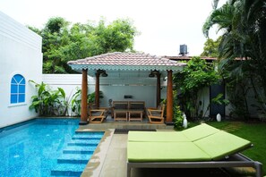 A pool area with a covered seating area, steps leading into the pool, and a lounge chair.