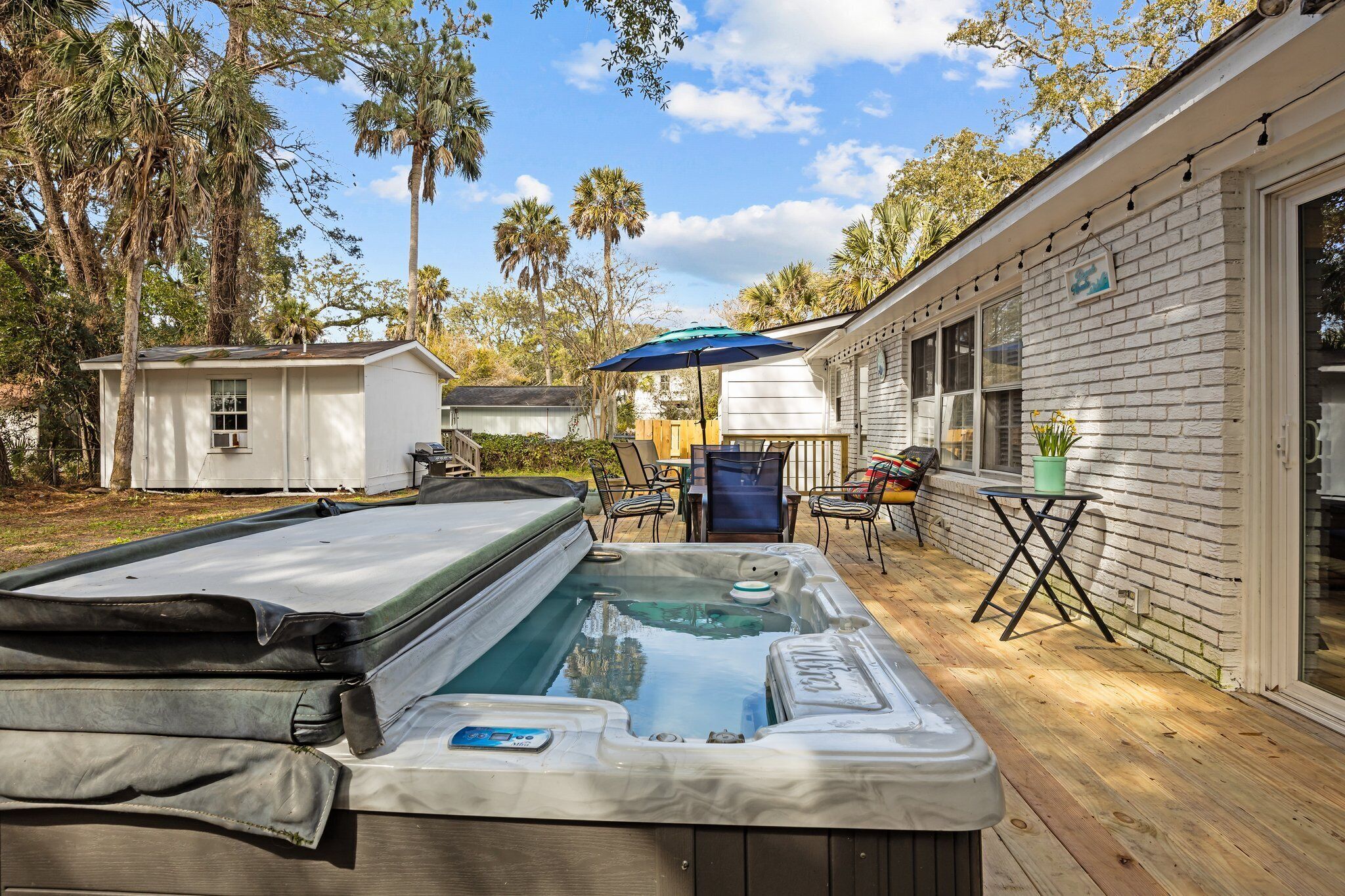 A hot tub on a wooden deck with a patio chair and umbrella, surrounded by a white building with large windows and a small shed in the background.