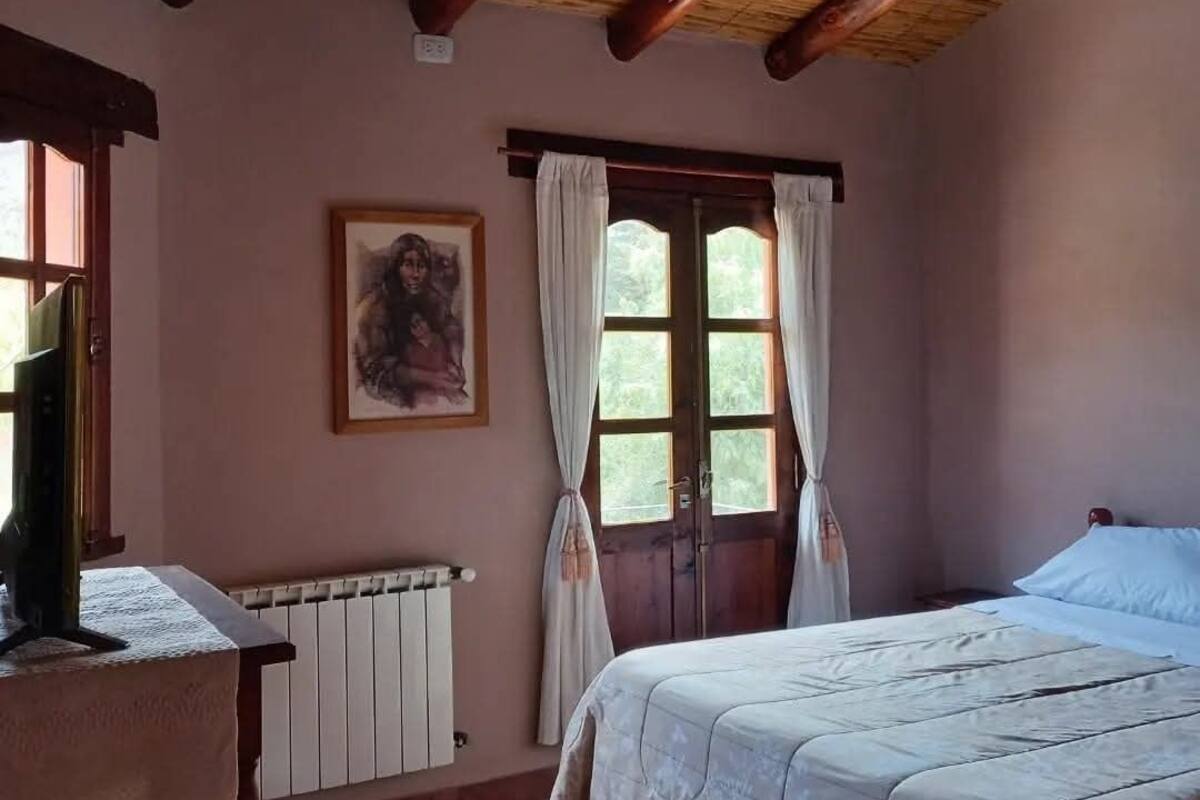 A bedroom with a wooden ceiling, a bed, a radiator, a window with curtains, and a painting on the wall.