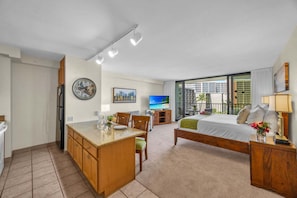 A compact hotel room with a kitchenette, a bed, a TV, and a balcony with a view.