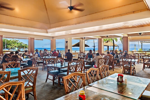 An outdoor dining area with wooden chairs and tables, large windows offering ocean views, and a ceiling fan.