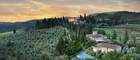 A villa with a swimming pool surrounded by olive groves and cypress trees, set on a hillside.