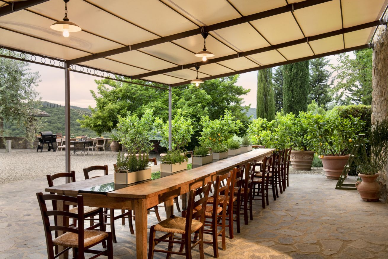 Een terras buiten met een lange houten tafel en stoelen, omringd door potplanten en een stenen vloer.