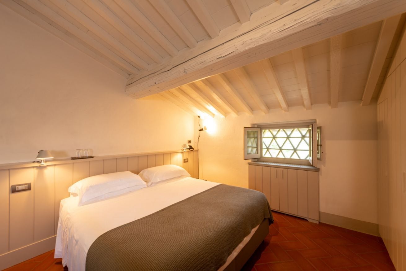 A bedroom with a bed, a nightstand, a wall-mounted lamp, and a window with a view.