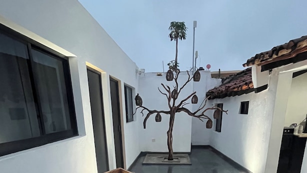 Un área de estar moderna al aire libre con dos mesas y sillas de madera, una escultura de árbol decorativa y piso de baldosas.
