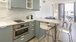 Una cocina moderna con horno empotrado, lavamanos y cafetera. Hay un comedor con mesa y sillas.