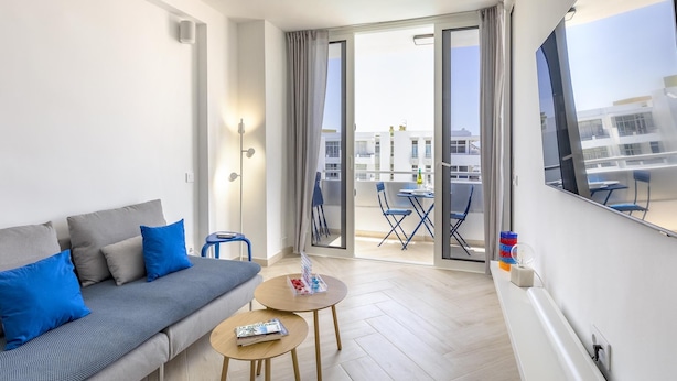 Una sala de estar moderna con un sofá gris, cojines azules, una mesita redonda y un balcón con sillas y una mesa azules.