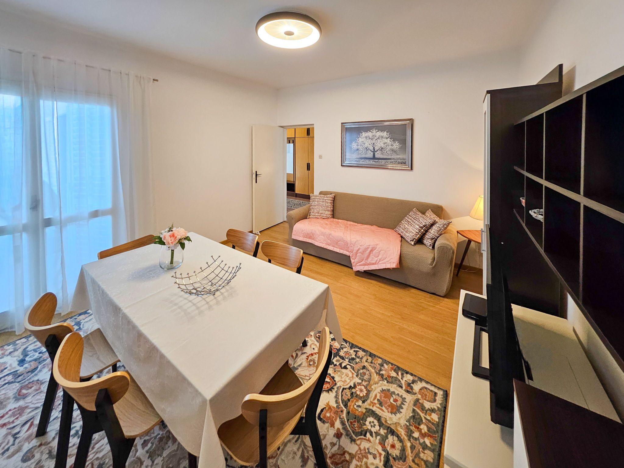 A dining area with a table and chairs, a sofa with a pink blanket, a framed picture on the wall, and a bookshelf.