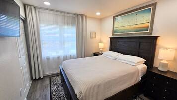 A hotel room with a bed, two bedside lamps, a painting of a beach scene, and a window with curtains.