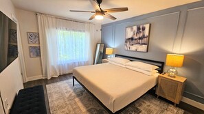 A bedroom with a bed, a ceiling fan, a nightstand with a lamp, a mirror, and a painting on the wall.